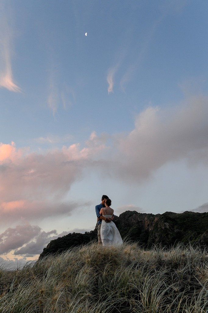 The couple stand against the enormous backdrop of a pink sunset