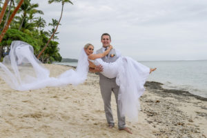 The groom carries his bride on the beach with her veil flowing behind them