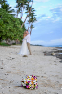 The camera focuses on the bouquet while the newly weds share a passionate kiss on Shangri La beach in the background